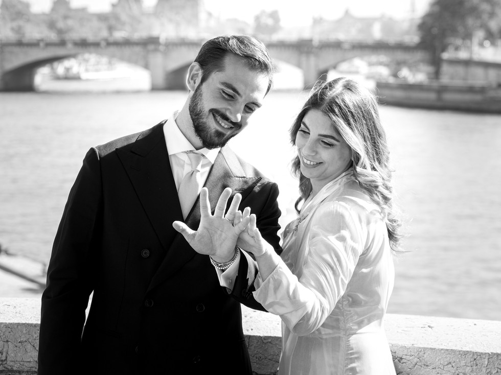 Photo en noir et blanc d’un couple de jeunes mariés souriants après leur mariage civil à Paris, admirant leurs alliances devant la Seine. Le marié porte un costume sombre et la mariée une robe fluide claire, baignée de lumière naturelle. Photographie signée Colombe Photographie, photographe de mariage civil à Paris.
