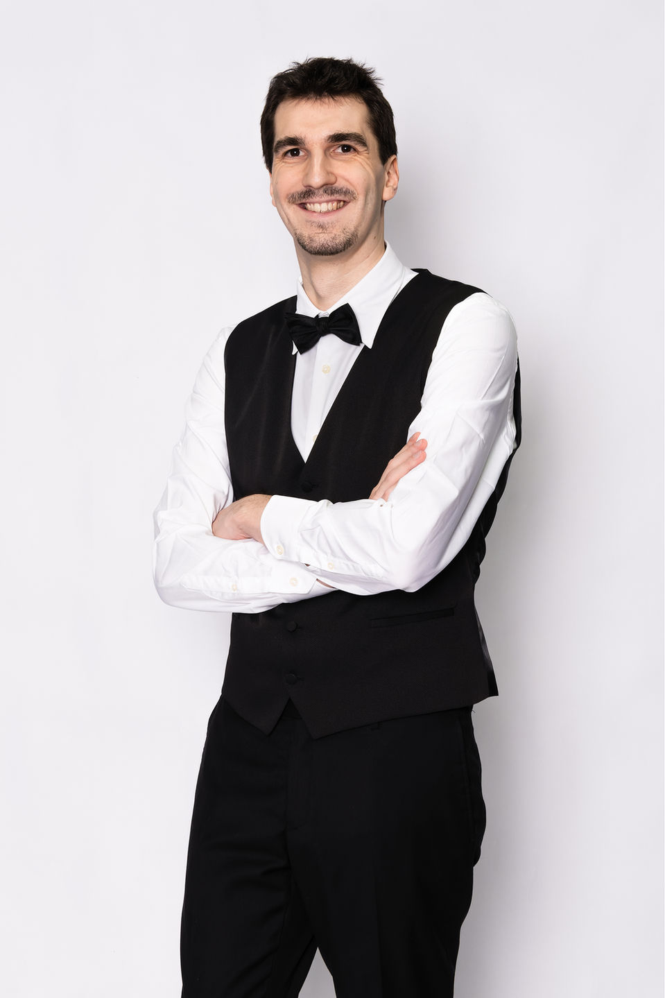 Pola de Raphaël Distler Stragier, comédien, en plan américain de trois quarts. Il pose avec les bras croisés, affichant un sourire naturel. Vêtu d'un pantalon, d'un gilet et d'un nœud papillon noir sur une chemise blanche, il incarne une élégance classique. Photo réalisée en studio sur fond blanc, sans retouche, idéale pour les agences et castings recherchant des profils authentiques et expressifs.
