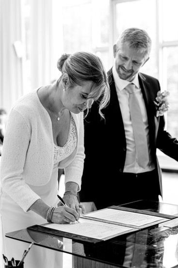 Photographie en noir et blanc de Camille signant les registres du mariage civil à la mairie du 17e arrondissement de Paris, sous le regard attendri de Xavier. Image capturée par Henock pour le studio Colombe Photographie, photographe de mariage civil à Paris et en région parisienne.