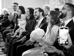 Photo en noir et blanc d’Annapaola et Angelo assis parmi leurs proches pendant leur mariage civil à la mairie du 7e arrondissement de Paris. Les mariés rient avec leurs familles, capturant un moment de joie et de complicité pendant la cérémonie. Cette image illustre la chaleur et l’émotion partagées lors de cette union, photographiée par Colombe Photographie, photographe de mariage civil à Paris et en région parisienne.