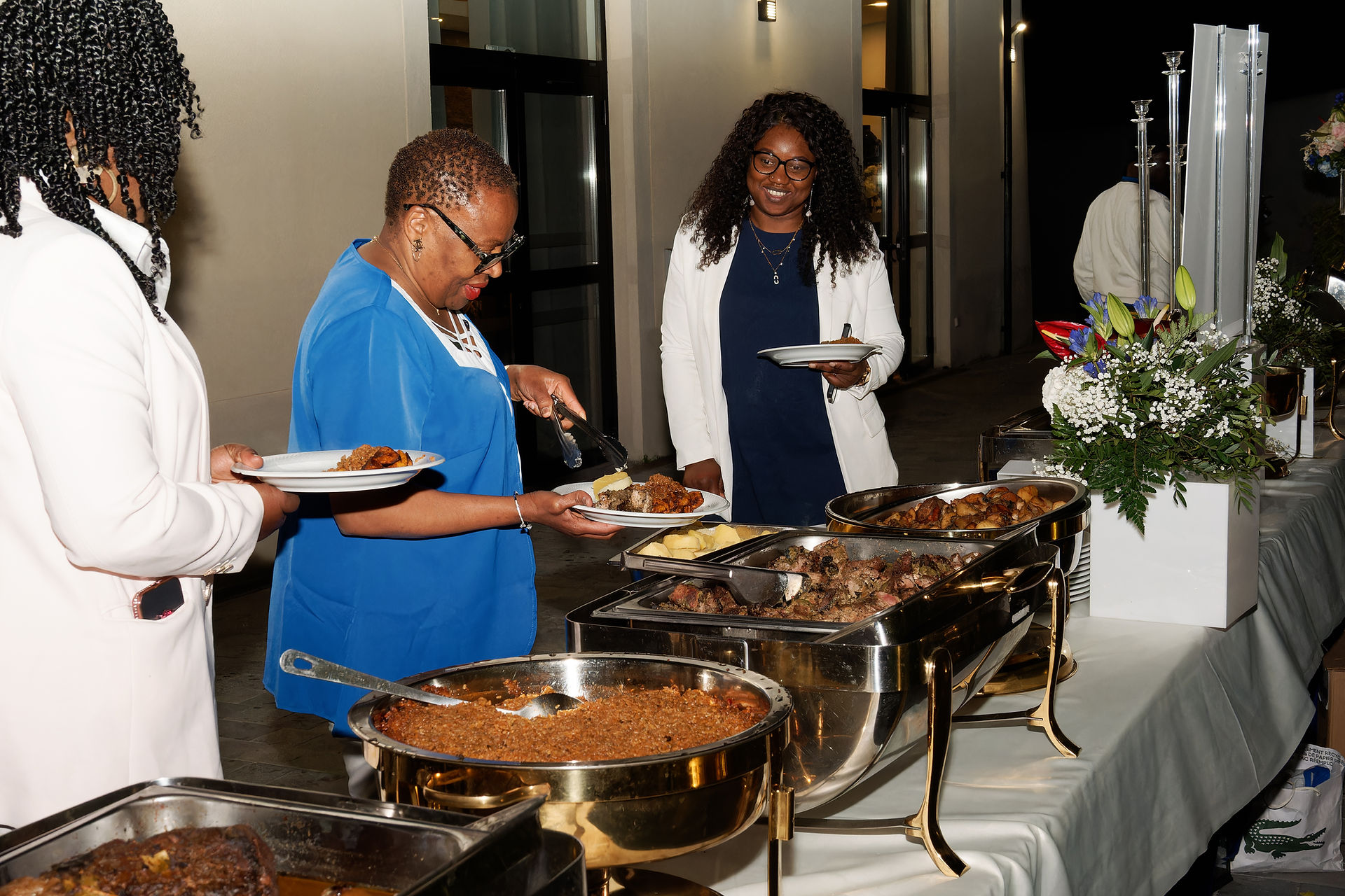 Photo prise pendant l’anniversaire surprise des 50 ans d’Armel, en région parisienne. Trois invitées se servent au buffet dans une ambiance conviviale en extérieur, devant une sélection de plats variés et colorés. Reportage réalisé par Colombe Photographie, photographe d’événements privés à Gentilly et en Île-de-France.