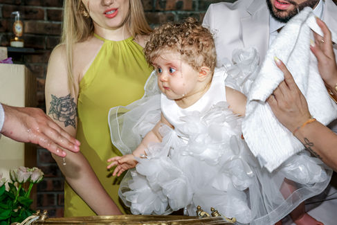 Bébé Giulia en robe blanche décorée de fleurs lors de son baptême à l’église Sainte Geneviève de Garges-lès-Gonesse. Les parents tiennent leur enfant au-dessus de la fontaine baptismale pendant que le prêtre verse l’eau bénite. Reportage réalisé par une photographe baptême à Paris et en Île-de-France, capturant l’émotion de la cérémonie familiale.