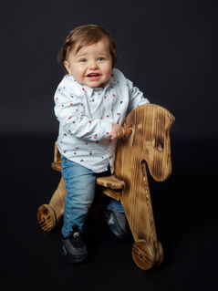 Portrait d’enfant réalisé au Studio Colombe Photographie à Gentilly, en région parisienne. Cette image met en scène un bébé souriant installé sur un cheval à bascule en bois, dans un décor studio simple et moderne. Le studio est spécialisé en photographie enfant et famille, offrant des portraits naturels, expressifs et chaleureux. Idéal pour une séance photo bébé ou famille en studio à Gentilly ou partout en Île-de-France.
