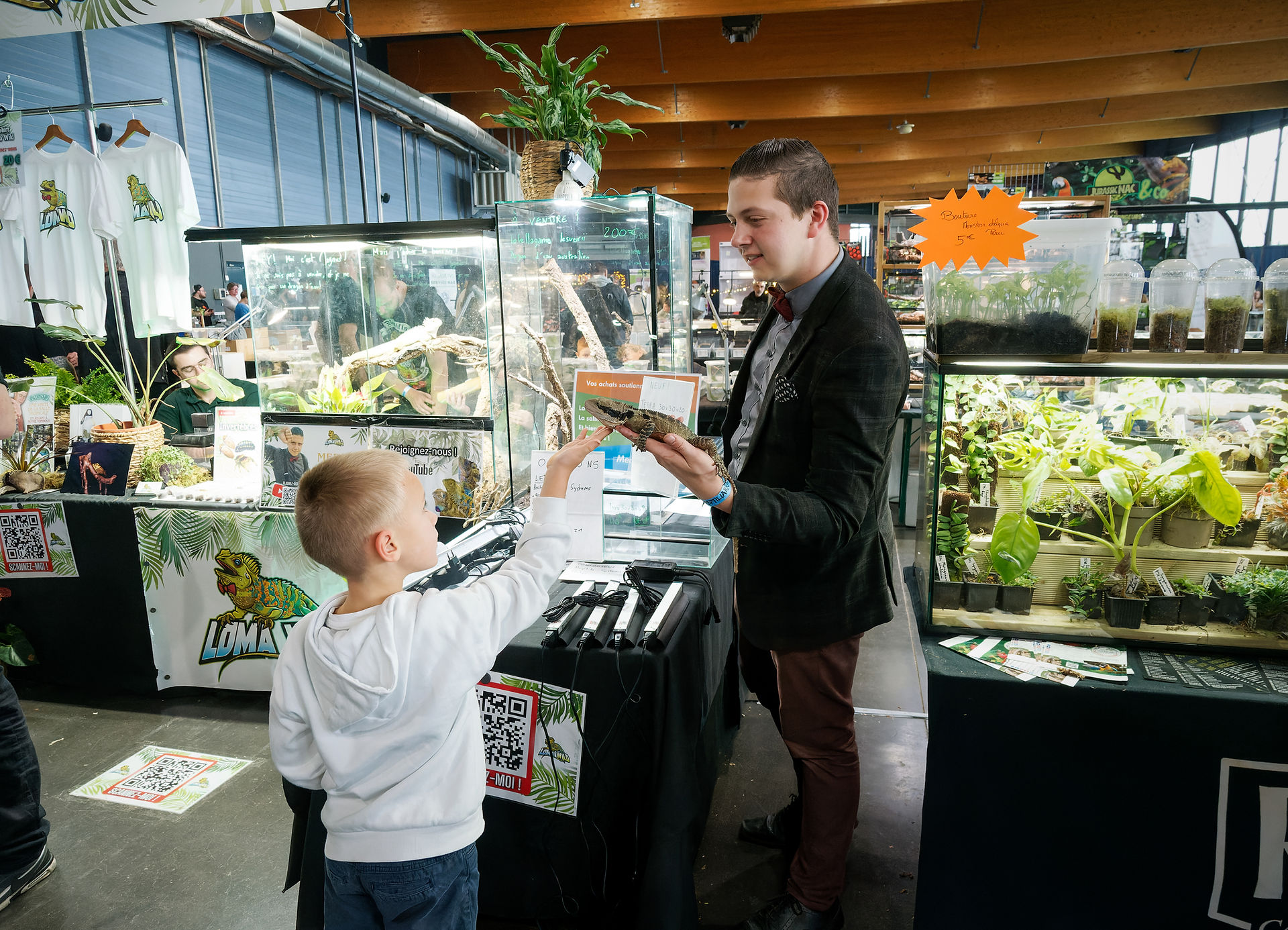 Scène de vie au stand Loma Wild lors des Reptiliarras 2025 : un visiteur enfant échange avec un exposant passionné devant les terrariums et les reptiles. La photo illustre l’aspect pédagogique et familial du salon. Réalisée par le studio Colombe Photographie, spécialisé en photographie événementielle et en reportage sur les expositions dédiées aux reptiles et arthropodes.