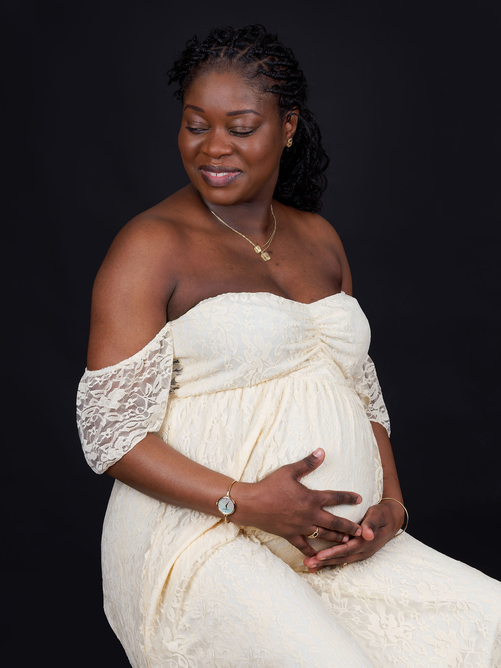 Ariane assise, mains sur le ventre, en robe de grossesse beige, regard tendre et souriante, photo grossesse en studio.