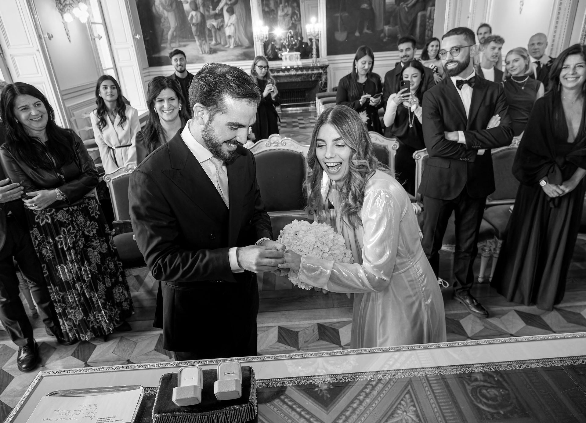 Photo en noir et blanc d’Annapaola et Angelo échangeant leurs alliances lors de leur mariage civil à la mairie du 7e arrondissement de Paris. Entourés de leurs proches, les mariés rient et partagent un moment de joie spontanée dans un décor élégant aux boiseries dorées. Ce cliché plein de vie et d’émotion a été capturé par Colombe Photographie, photographe de mariage civil à Paris et en région parisienne.