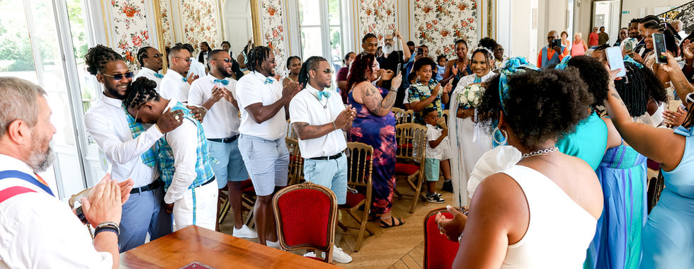 Photographie de l’arrivée de la mariée lors du mariage civil de Mallory et Ruddy, en Île-de-France. La salle est décorée de papiers peints fleuris et baignée de lumière naturelle. Entouré de ses proches, l’un des mariés laisse transparaître son émotion, soutenu par ses témoins. Photographie réalisée par Colombe Photographie, photographe de mariage à Gentilly et dans toute la région parisienne.