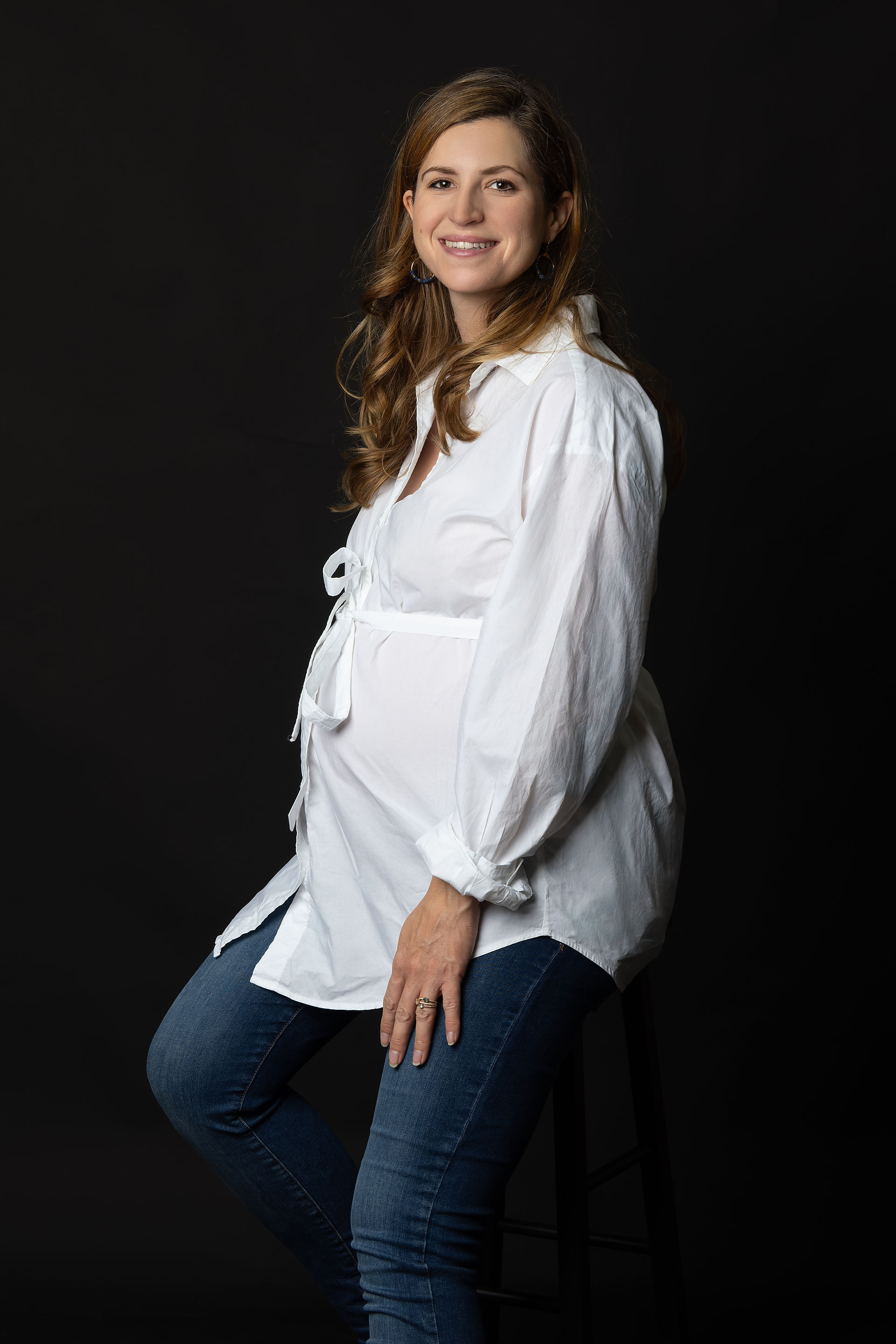 Portrait de femme enceinte seule en studio photo à Gentilly, près de Paris. Future maman assise sur tabouret, pose naturelle et élégante mettant en valeur le ventre arrondi. Séance photo grossesse réalisée par un studio photo en Île-de-France, idéale pour un souvenir intime et professionnel de la maternité.