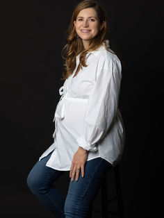 Amélie, assise sur un tabouret, de trois quarts. Son ventre rond est mis en valeur par une chemise blanche.