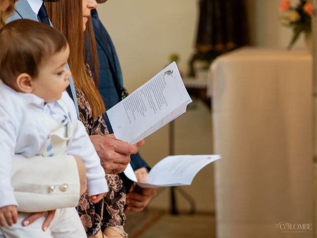 A religious baptism ceremony in the church, with a moment of liturgical chants. The baby, Samuel, is held in his mother's arms in the foreground. The soft light and the reverent atmosphere of a Christian baptism.