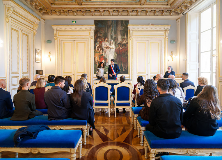 Vue d’ensemble de la salle des mariages du 7e arrondissement de Paris pendant le mariage civil d’Annapaola et Angelo. Les invités sont assis face à l’officier d’état civil et à la traductrice, dans un décor élégant aux boiseries dorées et aux sièges bleu royal. Cette image met en valeur la beauté du lieu et l’atmosphère solennelle de la cérémonie, photographiée par Colombe Photographie, photographe de mariage civil à Paris et en région parisienne.