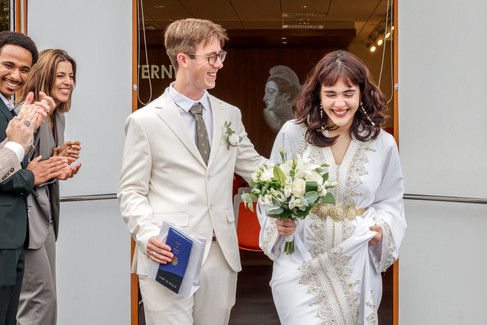 Photo de mariage civil à la mairie de Massy : Nathanaël et Hafsa, rayonnants de bonheur, sortent sous les applaudissements avec leur livret de famille en main. La mariée porte une robe traditionnelle blanche brodée et tient un bouquet de fleurs blanches. Photographe de mariage en Île-de-France, Colombe Photographie, basée à Gentilly, capture avec émotion les plus beaux instants.