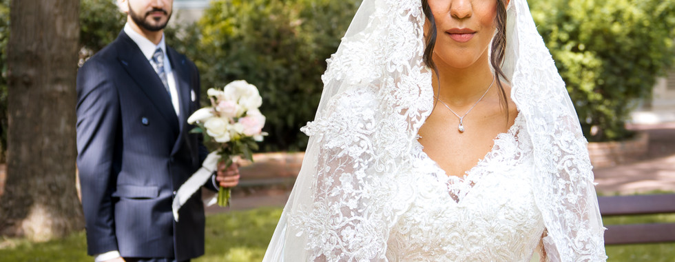 Photo de mariage d’Omer et Syrine en région parisienne : la mariée en robe blanche et voile dentelé, ceinte d’un ruban rouge, se tient au premier plan tandis que le marié s’avance derrière elle, un bouquet à la main.