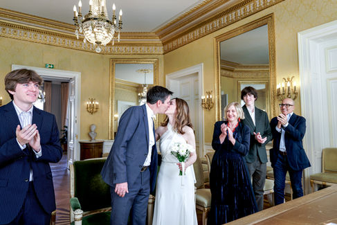 Photographe de mariage à Paris – moment fort du mariage civil de Daniela et David à la mairie du 9ème arrondissement. Les mariés s’embrassent après l’échange des vœux, sous les applaudissements de leur famille et amis. La mariée, en robe blanche fluide et bouquet à la main, et le marié, en costume sombre, sont entourés de sourires et d’émotions dans une salle dorée au décor raffiné. Ce cliché illustre toute l’intensité et la joie d’un mariage parisien capturé par Colombe Photographie, photographe de mariage en Île-de-France et Gentilly.