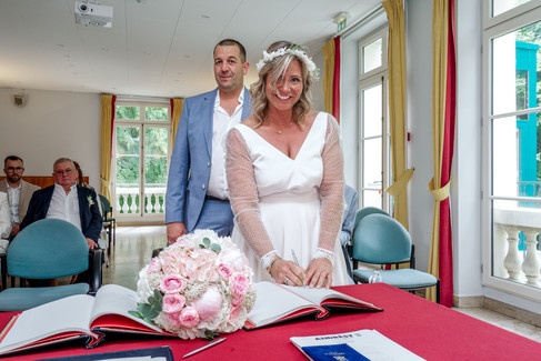 Photo de Mélanie signant le registre lors de son mariage civil avec Stéphane à la mairie de Conflans-Sainte-Honorine. La mariée rayonne de bonheur dans sa robe courte à manches en tulle plumetis, entourée de ses proches. Ce moment solennel et symbolique a été capturé par Colombe Photographie, photographe de mariage à Gentilly et en Île-de-France, spécialisée dans les cérémonies civiles intimistes et joyeuses.