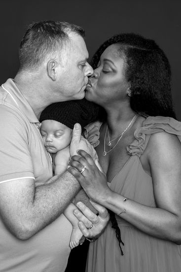 Photographie de famille capturant un instant tendre et spontané : les parents s’embrassent doucement tandis que leur bébé, Erwan, dort profondément dans les bras de son papa. Cette image illustre à la fois l’amour du couple et la sérénité du nourrisson, symbole de ce nouveau lien à trois. Un moment de complicité familiale, saisi avec délicatesse, dans une atmosphère chaleureuse et naturelle. Idéal pour immortaliser l’émotion des premiers jours de vie.