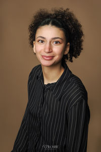 Portrait professionnel d'Ana, étudiante, réalisé en studio avec fond marron, plan américain. Elle regarde la caméra avec un sourire, habillée d'une chemise noire à rayures blanches, cheveux attachés. Cette image illustre les avantages d'une séance photo en studio, idéale pour des portraits destinés aux CV et profils professionnels, avec un éclairage contrôlé et un fond neutre qui met en valeur le sujet.