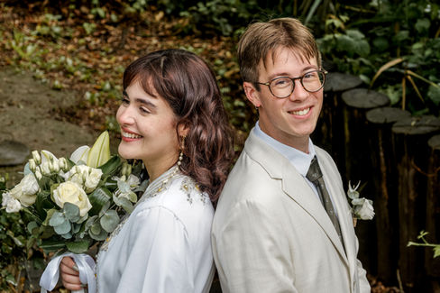 Portrait des mariés Nathanaël et Hafsa, pris juste après leur mariage civil à Massy. Debout dos à dos dans un cadre végétal, ils affichent de larges sourires complices. Hafsa tient un bouquet de fleurs blanches, vêtue d’une robe traditionnelle marocaine. Nathanaël porte un costume clair. Une photo signée Colombe Photographie, photographe de mariage à Gentilly et en Île-de-France, spécialisée dans les reportages authentiques et naturels.