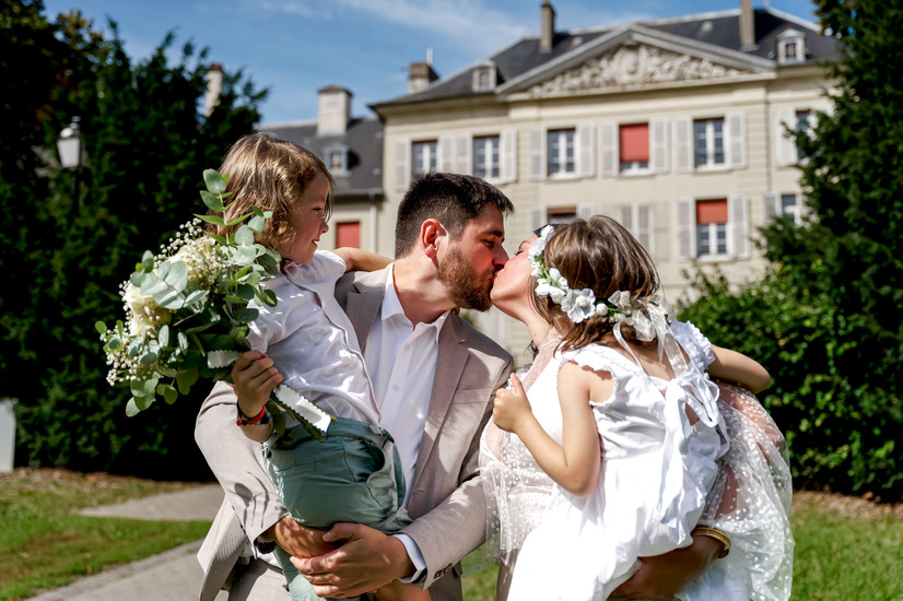 Photographie du mariage civil d’Élodie et Damien à Palaiseau : les mariés s’embrassent en tenant leurs enfants dans les bras, un instant de complicité familiale plein de joie et d’émotion. Reportage réalisé par Henock, photographe partenaire du Studio Colombe Photographie, spécialisé dans les mariages civils en Essonne et région parisienne.