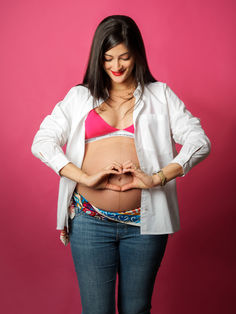 Femme enceinte sur fond rose formant un cœur avec ses mains autour de son ventre. Photographe grossesse à Gentilly en Île-de-France, spécialisé en portraits maternité créatifs et modernes en studio.