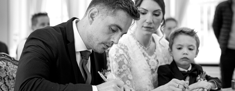 Photo en noir et blanc d’un mariage civil à Morsang-sur-Orge, montrant le marié signant les registres officiels, accompagné de sa femme et de leur jeune fils. Ce moment symbolique de la cérémonie illustre l’union familiale et la tendresse partagée. Image capturée par le Studio Colombe Photographie, photographe de mariage à Gentilly et en région parisienne.