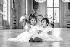 Photographie en noir et blanc réalisée lors du mariage civil de Chamima et Olivier à la mairie de Levallois-Perret. Deux enfants d’honneur, vêtus de jolies robes blanches, sont assis dans les salons d’honneur, souriants et complices, l’une tenant un bouquet de fleurs. Cette image illustre la spontanéité et l’émotion qui accompagnent les mariages en Île-de-France. Photographe de mariage à Levallois-Perret, je capture aussi bien les grands moments que les détails et les instants tendres qui donnent toute leur authenticité à votre journée.