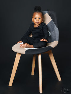 Portrait de Zeya, bébé célébrant ses 1 an, assise sur une chaise design en studio photo à Gentilly. Vêtue de noir sur fond sombre, elle regarde l’objectif avec douceur et curiosité. Photo réalisée par Colombe Photographie, photographe bébé et famille à Paris, spécialisée dans les séances anniversaires et portraits en lumière naturelle ou studio.