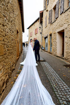 Photo de mariage de Rania et Jordan marchant dans une ruelle pittoresque aux maisons en pierre dorée, la longue traîne de la mariée déployée derrière elle. Photographie de Colombe Photographie, photographe de mariage à Lyon et en région parisienne.