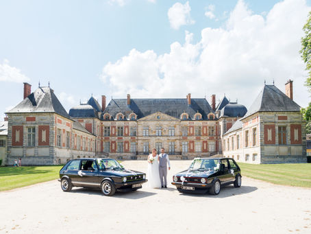 Photographie de mariage prise au Domaine de Courson en Essonne, montrant les mariés Audrey et Manu posant devant la façade majestueuse du château, entourés de deux voitures anciennes décorées pour l’occasion. Cette image symbolise l’élégance et le caractère historique du lieu de réception choisi pour leur mariage. Reportage réalisé par une photographe professionnelle spécialisée dans les mariages en Essonne et en Île-de-France.
