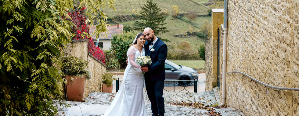 Photo de mariage de Rania et Jordan dans une ruelle pavée du Beaujolais, entre murs en pierre dorée et paysages de vignes. La mariée porte une robe blanche à longues manches perlées, le marié un costume bleu marine. Photographie de couple signée Colombe Photographie, photographe de mariage à Lyon et en région Auvergne-Rhône-Alpes.