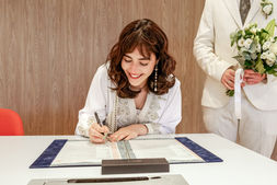 Photographie de Hafsa, la mariée, en train de signer le registre officiel lors de son mariage civil avec Nathanaël à la mairie de Massy. Elle porte une robe traditionnelle blanche brodée de perles et de dorures, et sourit avec émotion. Le marié, vêtu d’un costume ivoire, tient son bouquet à ses côtés. Reportage réalisé par Colombe Photographie, photographe de mariage civil à Gentilly et en Île-de-France.