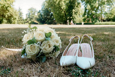 Photographie de détail capturée lors du mariage de Mallory & Ruddy en Île-de-France, montrant un bouquet de mariée composé de roses blanches et de gypsophile, posé à côté d'une paire d'escarpins blancs sur une pelouse ensoleillée. Ce cliché en extérieur reflète l’ambiance champêtre et raffinée du jour J. Prise de vue réalisée par Colombe Photographie, photographe de mariage basée à Gentilly et active dans toute la région parisienne.