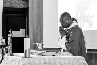 Photographie en noir et blanc d’un prêtre en prière devant l’autel pendant une messe de première communion, mains jointes et yeux fermés, dans une église baignée de lumière. Cette image a été prise à la paroisse Notre-Dame du Sacré-Cœur de Coeuilly. Photographe professionnelle d’événements religieux à Gentilly et en Île-de-France, je saisis l’émotion et la spiritualité de vos célébrations.