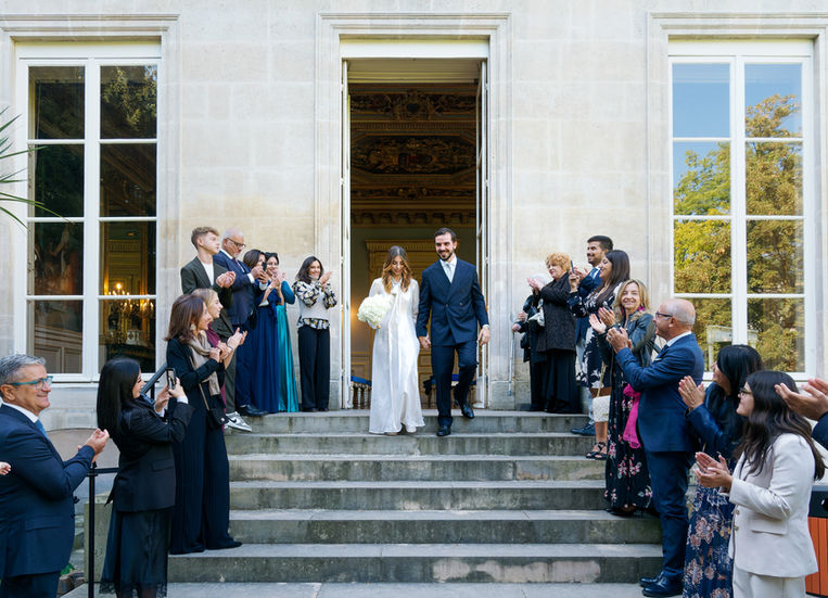 Photo couleur d’Annapaola et Angelo descendant les marches de la mairie du 7e arrondissement de Paris après leur mariage civil. Les mariés, main dans la main, sont entourés de leurs invités qui les applaudissent avec émotion à la sortie de la cérémonie. Une scène lumineuse et joyeuse capturée par Colombe Photographie, photographe de mariage civil à Paris et en région parisienne.