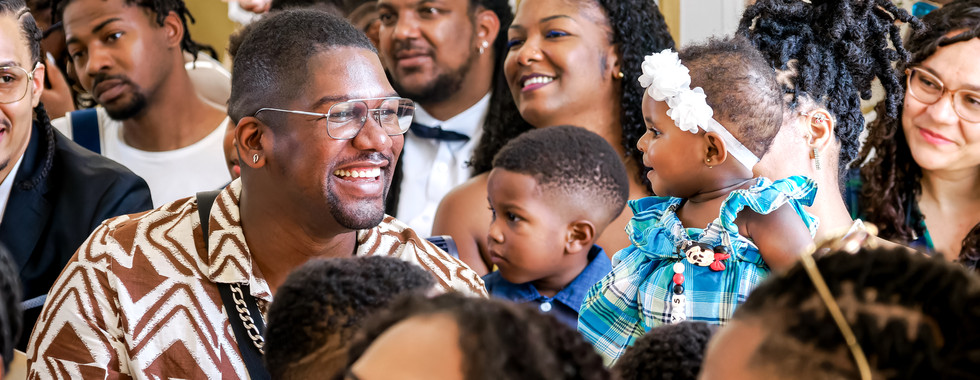 Photographie prise lors du mariage civil de Mallory et Ruddy à Gentilly, montrant un moment de joie parmi les invités. Un homme éclate de rire tandis qu’une petite fille en robe à carreaux madras est portée dans les bras, tournant le regard vers lui. Ce cliché illustre l’ambiance chaleureuse et familiale de la cérémonie, captée par Colombe Photographie, photographe de mariage en région parisienne et spécialisée dans les reportages naturels et pleins de vie.