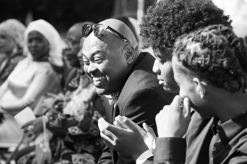 Photographie en noir et blanc d’invités riant pendant la cérémonie du mariage de Malika et Richard à Guyancourt, en Île-de-France. Reportage mariage plein de vie et d’émotion réalisé par Colombe Delons, photographe de mariage en région parisienne.
