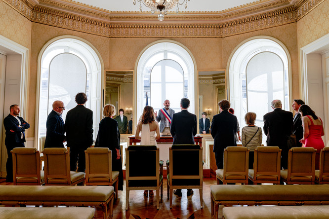 Photographe de mariage à Paris – reportage du mariage civil de Daniela et David à la mairie du 9ème arrondissement. Les mariés, vus de dos, se tiennent devant l’officier d’état civil portant l’écharpe tricolore. Autour d’eux, famille et amis proches assistent à la cérémonie dans une salle élégante aux dorures raffinées. Ce cliché illustre l’ambiance solennelle et intime d’un mariage en petit comité à Paris, immortalisé par Colombe Photographie, photographe de mariage en région parisienne et en Île-de-France.