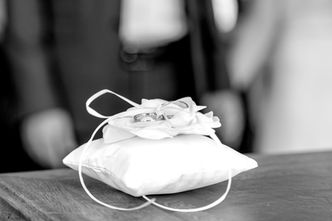 Photographe de mariage à Paris – détail des alliances de Daniela et David posées sur un coussin blanc, prêtes pour l’échange des vœux à la mairie du 9ème arrondissement. Ce cliché en noir et blanc capture l’élégance et la symbolique du mariage civil, avec le coussin orné d’un ruban et d’une fleur en tissu. Un instant délicat et intemporel qui illustre l’importance des détails dans un reportage de mariage. Colombe Photographie, photographe de mariage en région parisienne, Gentilly et Île-de-France, immortalise avec sensibilité chaque moment clé.