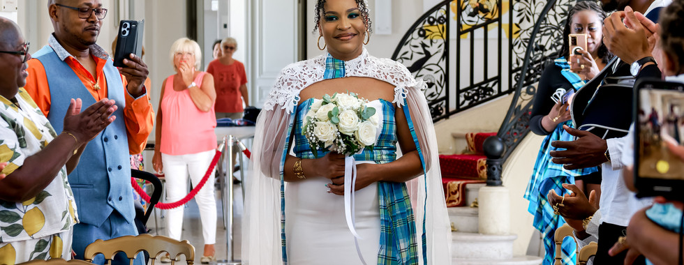 Photographie de l'entrée de la mariée lors du mariage civil de Mallory et Ruddy, en Île-de-France. Vêtue d'une robe blanche et d'une étole en dentelle avec une touche de madras bleu, elle avance souriante, un bouquet de roses blanches à la main. Capturée par Colombe Photographie, photographe de mariage basée à Gentilly et active dans toute la région parisienne.