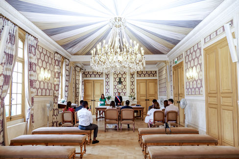 Photo de la cérémonie de mariage civil de Chamima et Olivier à la mairie de Levallois-Perret, capturée par Colombe Photographie. L’image montre les mariés de dos face à l’élu·e et leurs invités, dans une élégante salle ornée d’un lustre en cristal et de papiers peints floraux. Photographe de mariage en Île-de-France, basée à Gentilly et spécialisée dans les mariages en mairie en région parisienne.