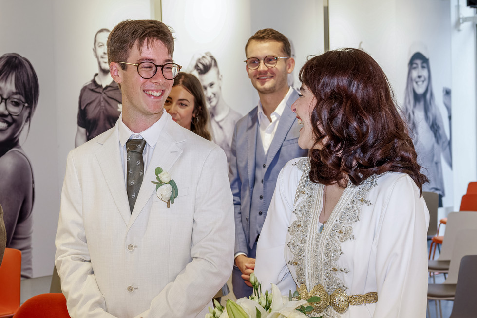 Photographie prise lors du mariage civil de Nathanaël et Hafsa à la mairie de Massy, en Île-de-France. On y voit les mariés échanger un regard complice, souriants et émus au cœur de la cérémonie. La mariée porte une tenue traditionnelle ornée de broderies argentées, le marié un costume blanc cassé avec une boutonnière florale. Reportage signé Colombe Photographie, photographe de mariage civil à Gentilly et en région parisienne.