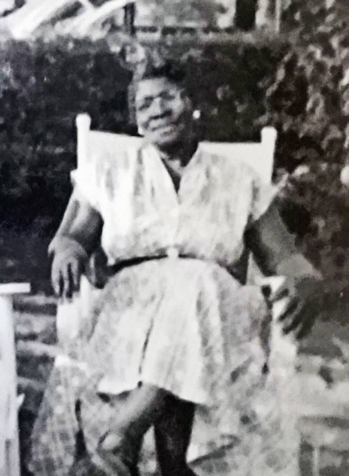Woman in a patterned dress sits on a white rocking chair, smiling in a garden setting. Black and white photo, with lush greenery behind her.