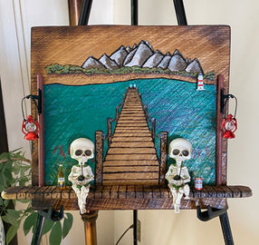 The hand-painted scene features a rustic dock extending into vibrant blue waters, leading toward majestic mountains. Two playful skeleton figurines sit at the dock’s edge, each holding a drink, symbolizing the importance of relaxation, laughter, and seizing the moment.