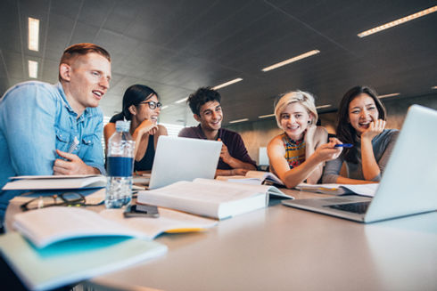 70199366-university-students-sitting-together-at-table-with-books-and-looking-at-laptop-ha