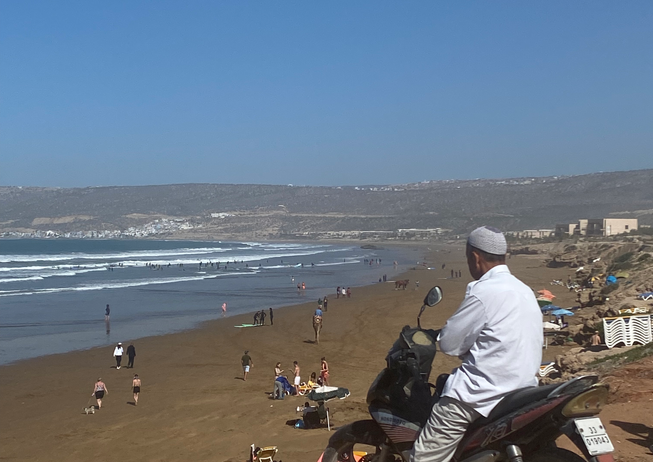 Un monsieur assis sur sa moto regarde la plage qui se situe plus loin et plus bas.