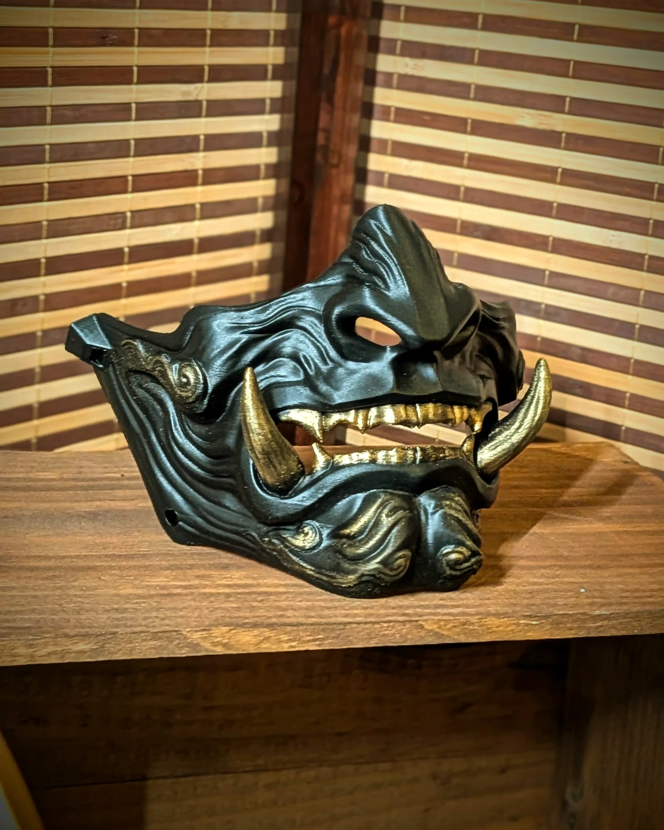 Handmade black Mempo Oni half-mask in PETG by Dai Yokai, with gold fangs and a dark patina, inspired by samurai war armor