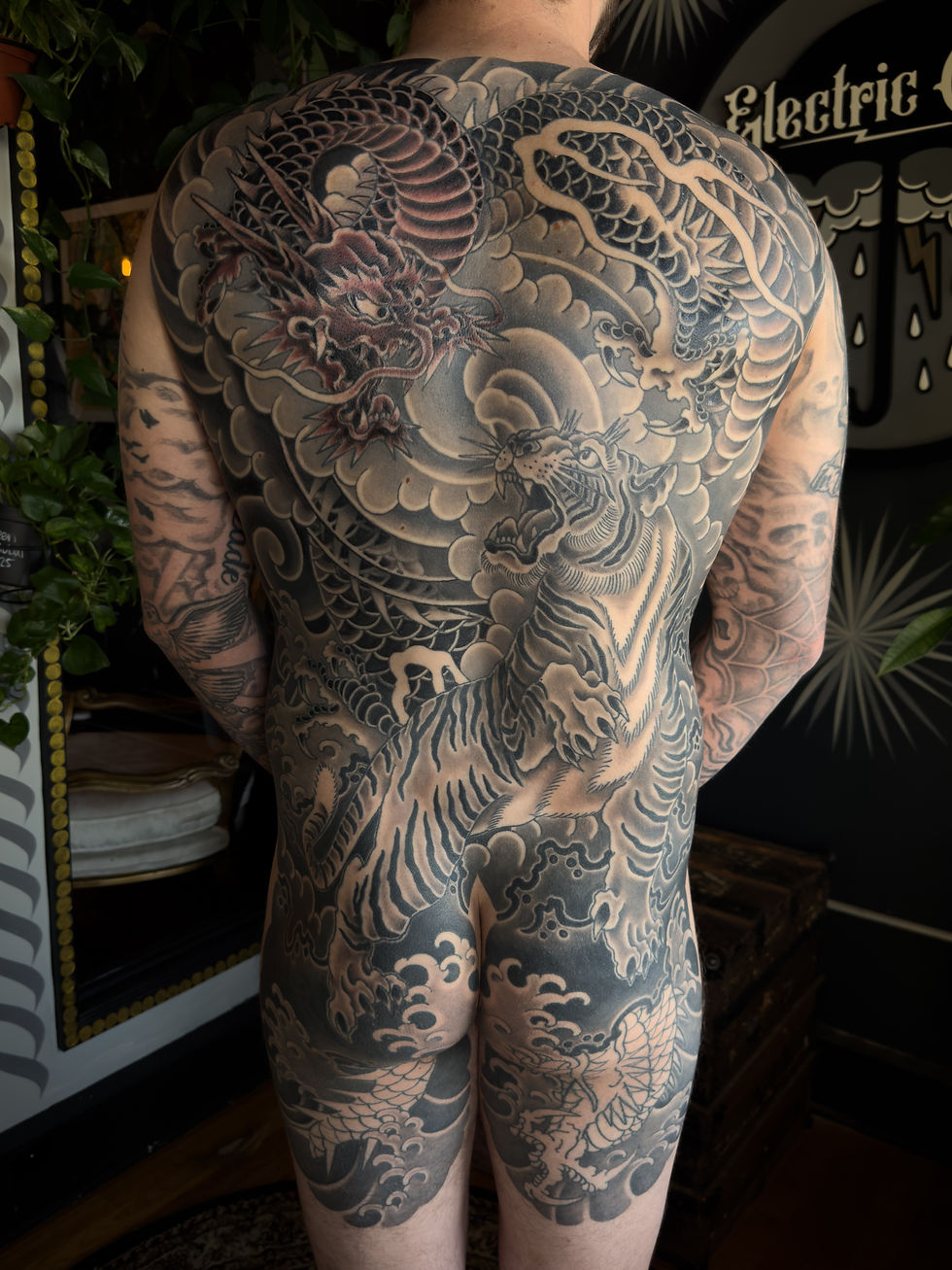 Man with full-back tattoo of dragon and tiger in grayscale. Tattoo studio backdrop with plants.