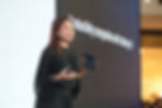 Woman in black presenting on stage, holding a dark tablet. Text on screen reads "Productivity companion with Galaxy." Bright, formal setting.