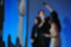 Three people pose for a selfie on stage with microphones. A woman in a glittery dress holds a tablet. Blue background and projection screen.