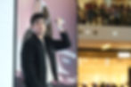 Man in black jacket presents a white smartphone on stage. Large screen displays a watch. Audience visible in background, indoor setting.